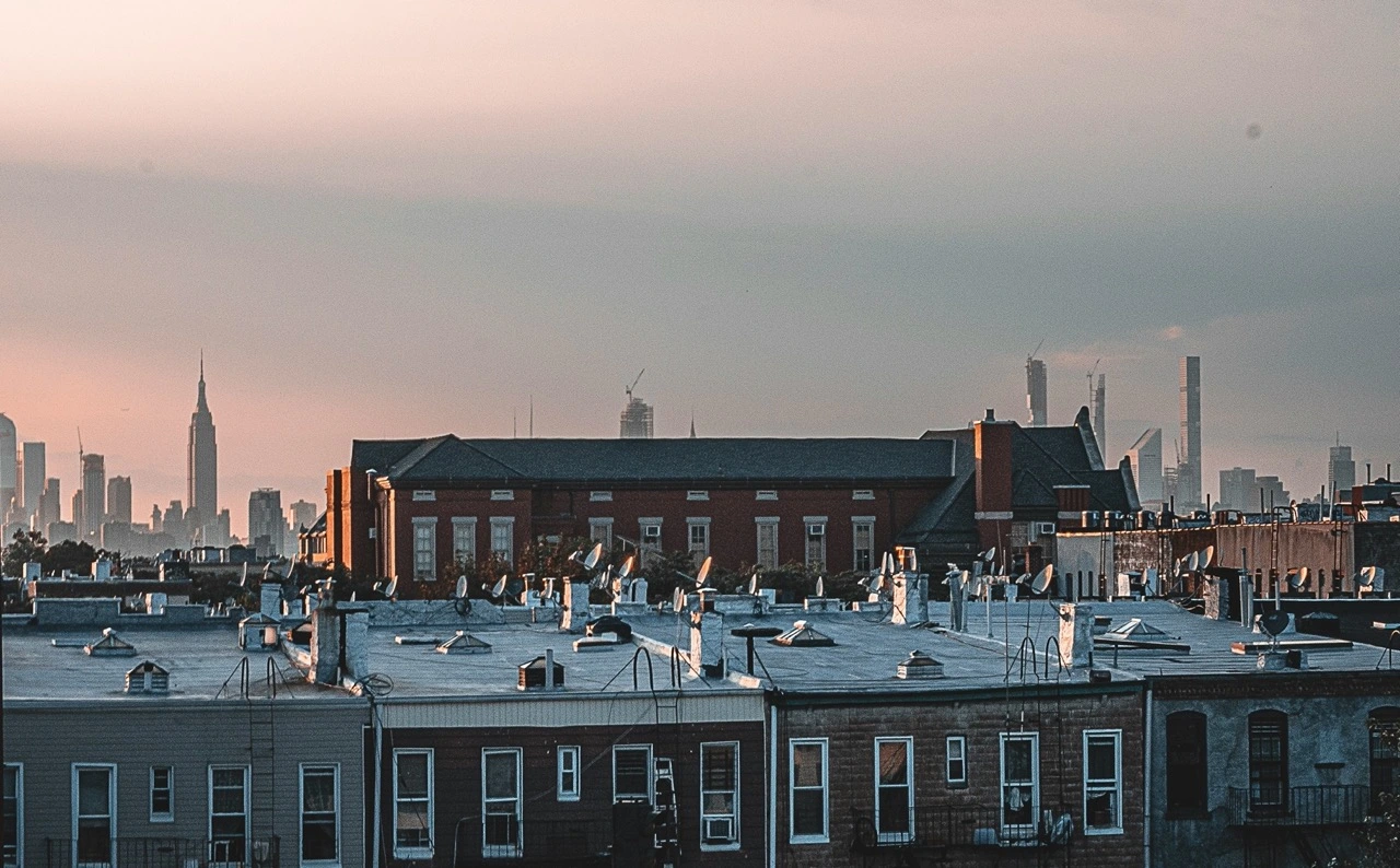 A New York City rooftop at dusk, looking out over Brooklyn's low-rise brick buildings toward the elevated subway tracks, the city grid receding into the haze.